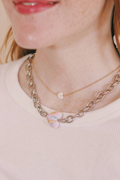 A woman wears the Dainty Mother of Pearl Layer necklace— a 14k gold filled chain with a small mother of pearl heart charm— layered with a chunky silver chain and pastel multicolored heart pendant, over a white textured top.