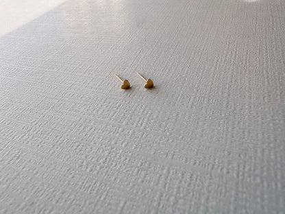 The Mini Heart Studs, crafted in 14k gold fill, are shown side by side on a textured white surface with a subtle grid pattern. The softly lit background highlights the delicate design.