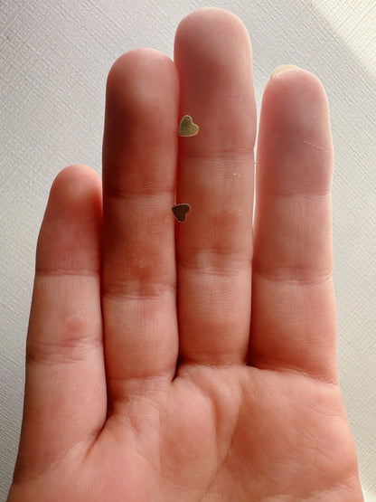 A close-up shows a hand with two Mini Heart Studs, delicate 14k gold filled heart-shaped earrings, resting on the middle and ring fingers against a light textured background.