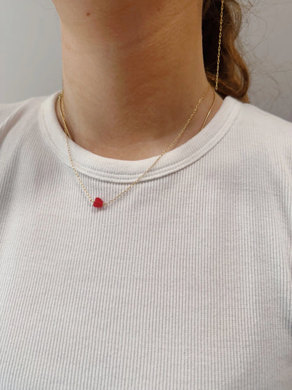 A person wears a white ribbed shirt and The Boyfriend Necklace, a delicate gold filled chain with a small red bead pendant; a thin gold filled earring is partially visible on one ear.