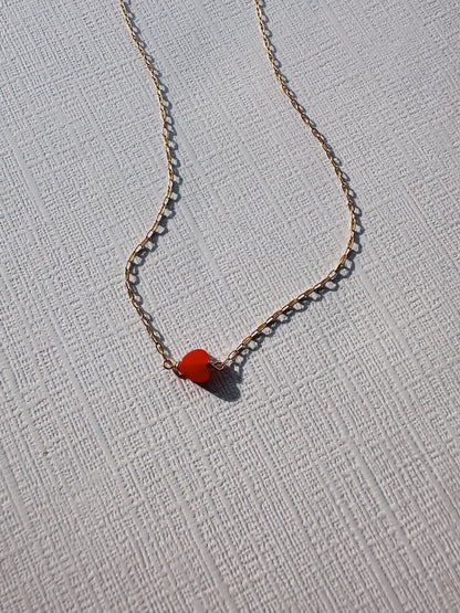 The Boyfriend Necklace, a delicate gold-filled chain with a small red heart-shaped gemstone pendant, is displayed on a textured white surface under soft natural light.