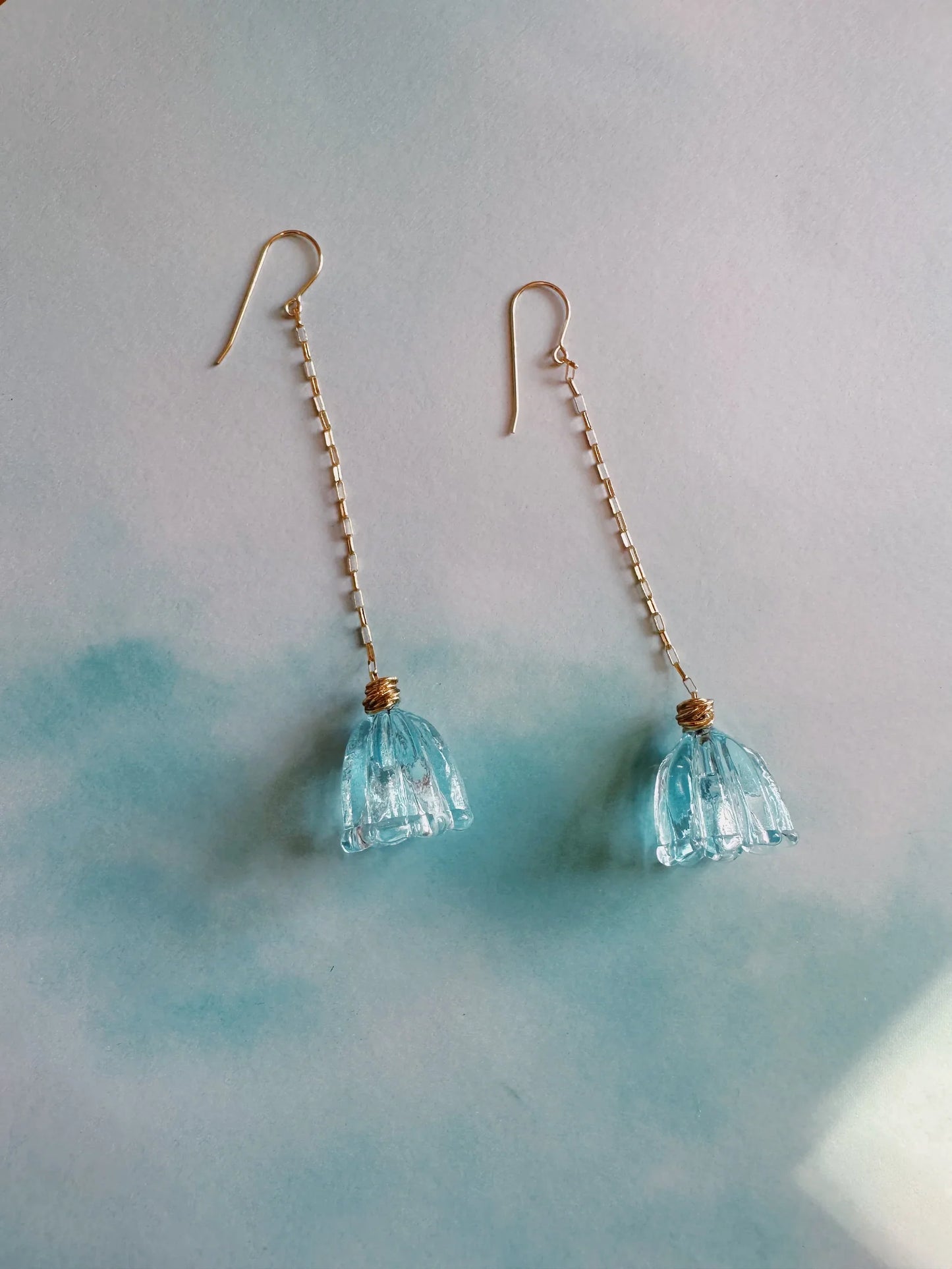 Glass Tulip Earrings in Clear Aqua feature long, delicate, artisan-crafted flower pendants on 14k gold-filled chain hooks, shown on a soft light blue and white background.