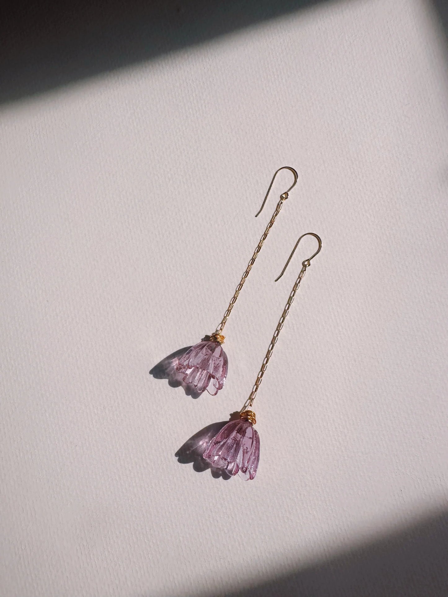 The Glass Tulip Earrings feature long 14k gold-filled chains with delicate, handmade magenta glass tulips at the ends, shown on a textured white surface with gentle light and shadow for an elegant look.