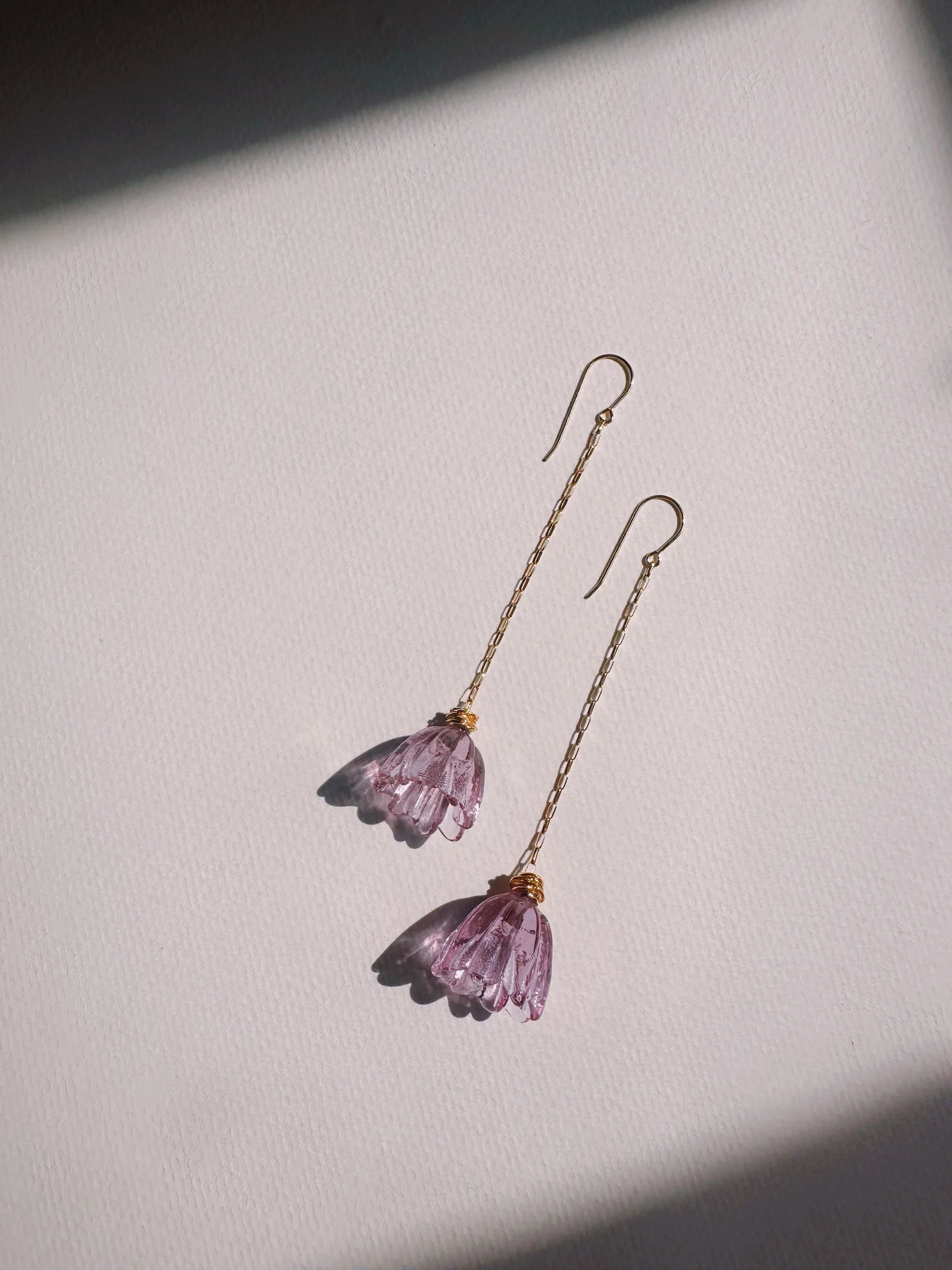 The Glass Tulip Earrings feature long 14k gold-filled chains with delicate, handmade magenta glass tulips at the ends, shown on a textured white surface with gentle light and shadow for an elegant look.