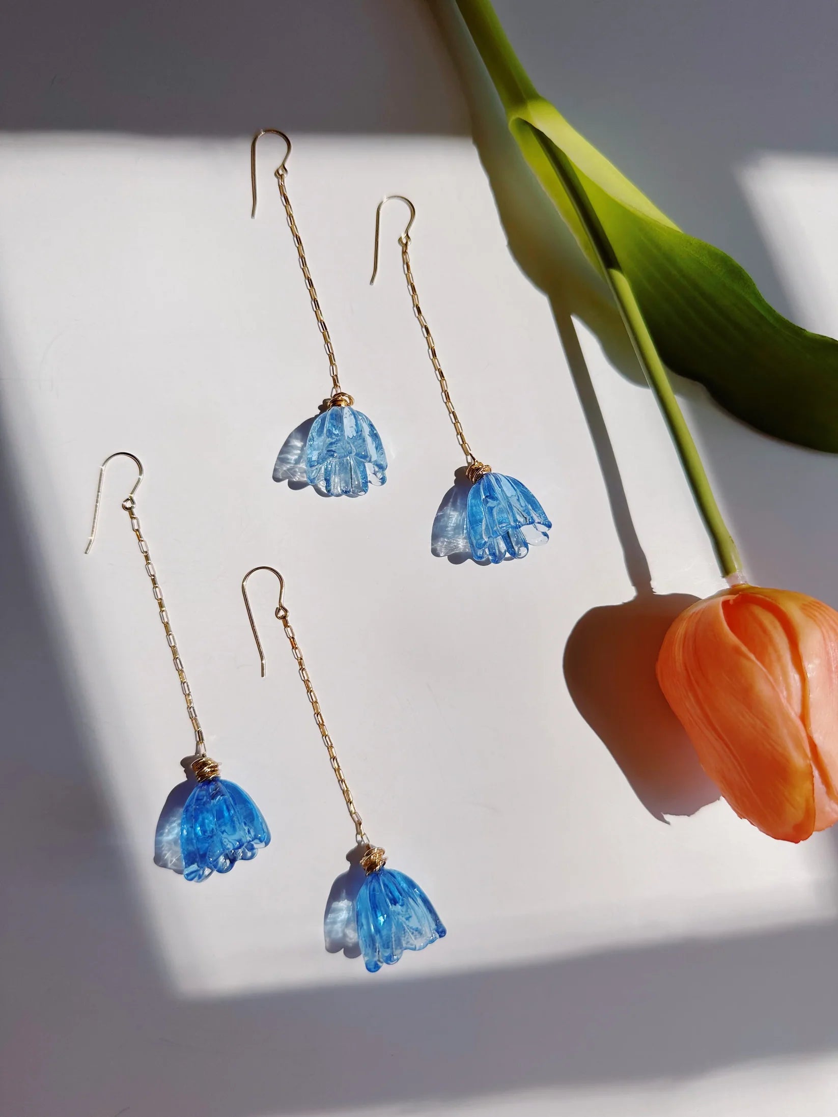 Five Glass Tulip Earrings in Marina Blue, with 14k gold-filled chains and flower-shaped pendants, are arranged on a white surface beside an orange handmade glass tulip with a green stem, illuminated by natural sunlight.