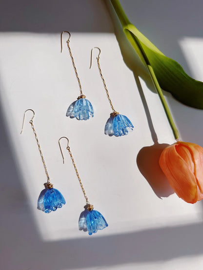 Five Glass Tulip Earrings in Marina Blue, with 14k gold-filled chains and flower-shaped pendants, are arranged on a white surface beside an orange handmade glass tulip with a green stem, illuminated by natural sunlight.