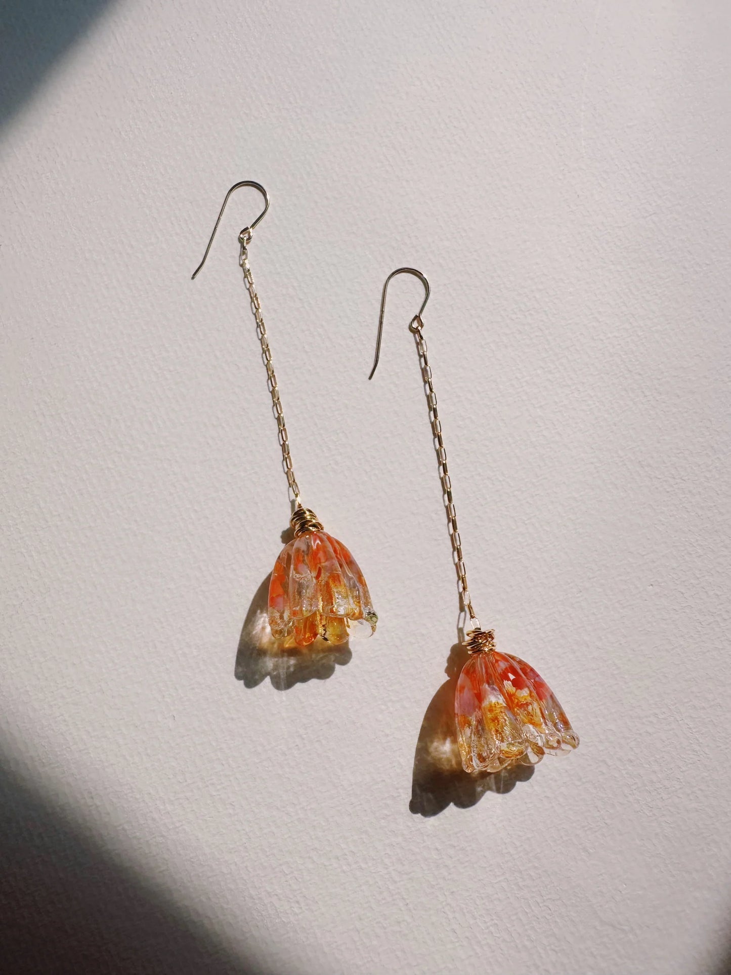 Glass Tulip Earrings: Delicate gold earrings with long 14k gold-filled chains, each featuring a speckled orange handmade glass tulip, displayed on a white textured surface in soft natural light.