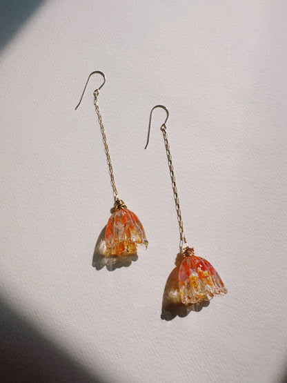 Glass Tulip Earrings: Delicate gold earrings with long 14k gold-filled chains, each featuring a speckled orange handmade glass tulip, displayed on a white textured surface in soft natural light.