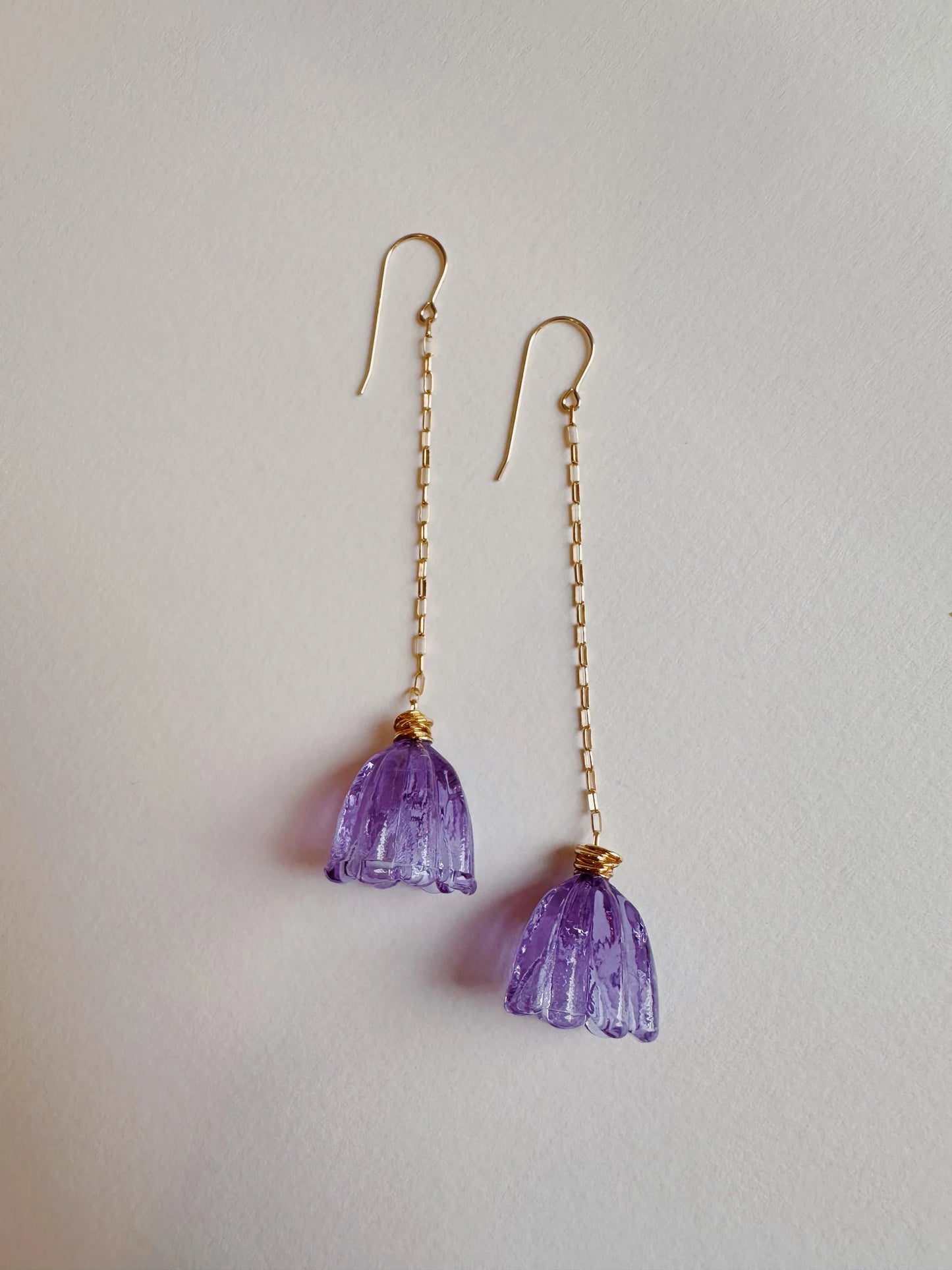 The Glass Tulip Earrings feature dangling, translucent violet glass tulip pendants handcrafted in a flower shape, suspended from a 14k gold-filled chain and shown on a plain white background.