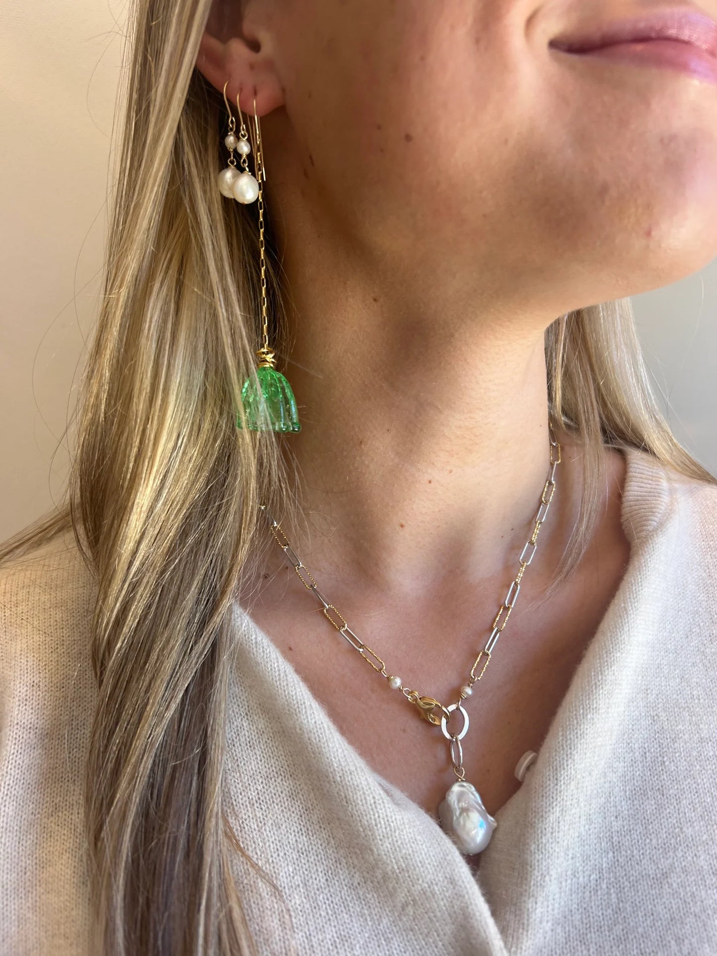 A woman with long blonde hair wears artisan jewelry: a pearl drop earring, handmade Glass Tulip Earrings in sea green, and a gold chain necklace with a large baroque pearl pendant. She is dressed in a light-colored top.